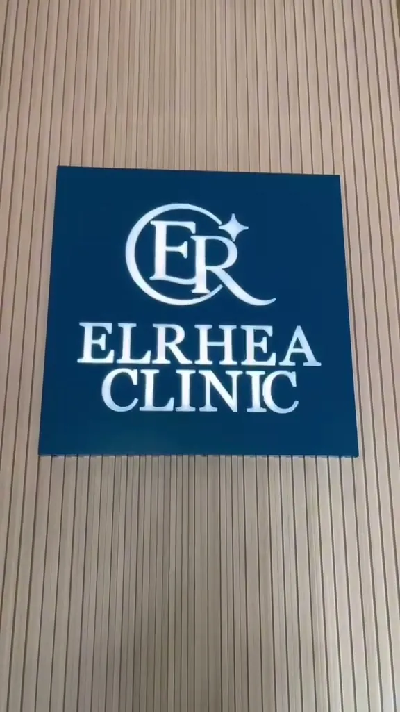 Papan nama Elrhea Clinic warna biru di dinding kayu